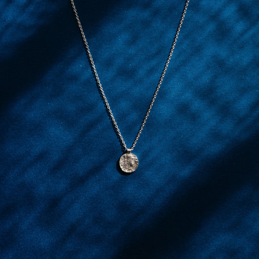A silver coin pendant necklace on a delicate silver chain, displayed against a textured royal blue fabric background with soft leaf shadows.