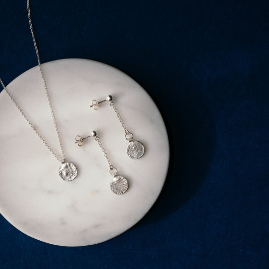 Silver jewelry set featuring a necklace and matching dangle earrings with textured circular disc pendants, displayed on a round white marble tray against a dark blue background.