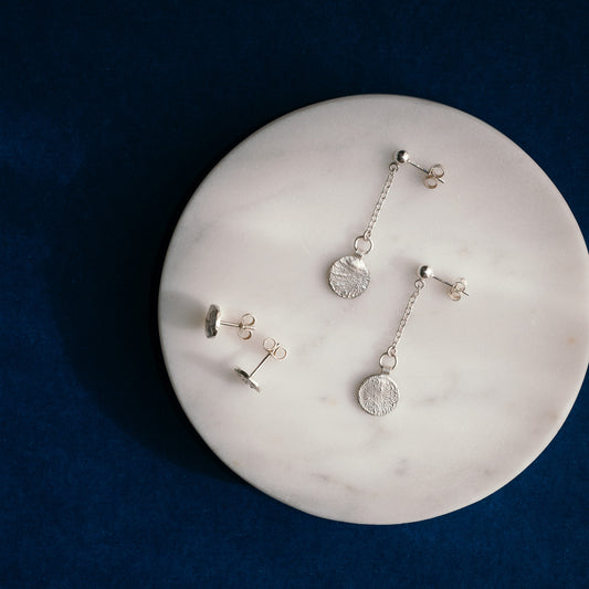 Three pairs of silver dangle earrings with textured circular disc charms and chain links, displayed on a round white marble tray over a dark blue background.