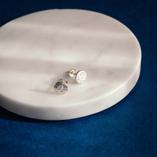A pair of small silver stud earrings with textured, hammered circular faces, resting on a round white marble tray against a dark blue velvet background.