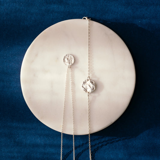 A matching pair of silver jewellery. A silver pendant necklaces with textured circular disc hanging from a fine chain, and a delicate silver chain bracelet with a textured circular medallion pendant, displayed on a round white marble coaster against a dark blue velvet background.