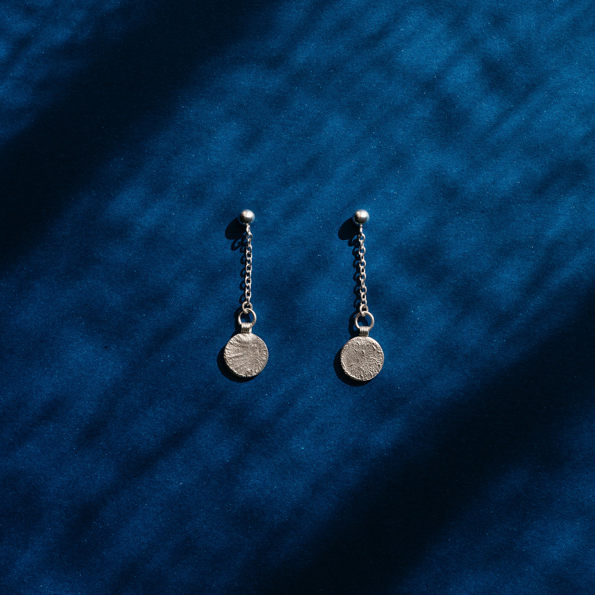 A pair of silver dangle earrings with small chain links and round coin-style pendants, displayed on a deep blue textured fabric background with soft shadow overlays.