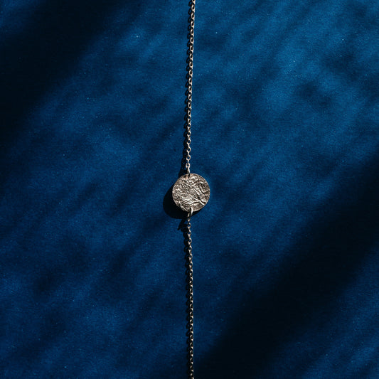 A silver chain necklace with a small, textured circular medallion pendant, laid flat on a deep blue velvet background with soft diagonal shadows.