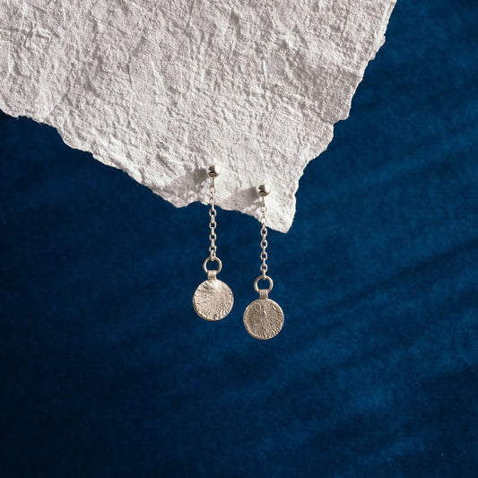 A pair of silver dangle earrings with textured circular coin pendants, hanging from delicate chains. They are displayed against a dark blue velvet background with a white textured stone prop at the top.