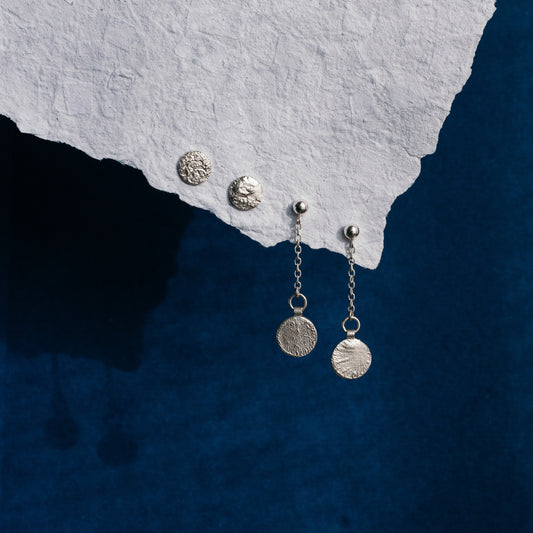 Two pairs of silver earrings displayed on a textured white paper corner against a deep blue background.  A pair of dangle earrings with textured coin-like discs hanging by chain from silver balls, and matching textured disc stud earring rests on the paper.